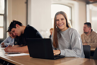 Several young adults sitting in a classroom. Young woman in the foreground working on a laptop looks smiling into the camera.