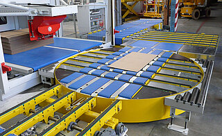 In the centre oft he picture there is a turntable with plastic chain conveyor and angular transfer. In the foreground there is a chain conveyor with 4 chain belts, in the background a angular transfer unit and a plastic chain conveyor. On the left you can see a further plastic chain conveyor.