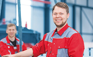 Ein Arbeiter von MINDA im Vordergrund mit einem roten Arbeitsanzug mit grauen Elementen lächelt in die Kamera